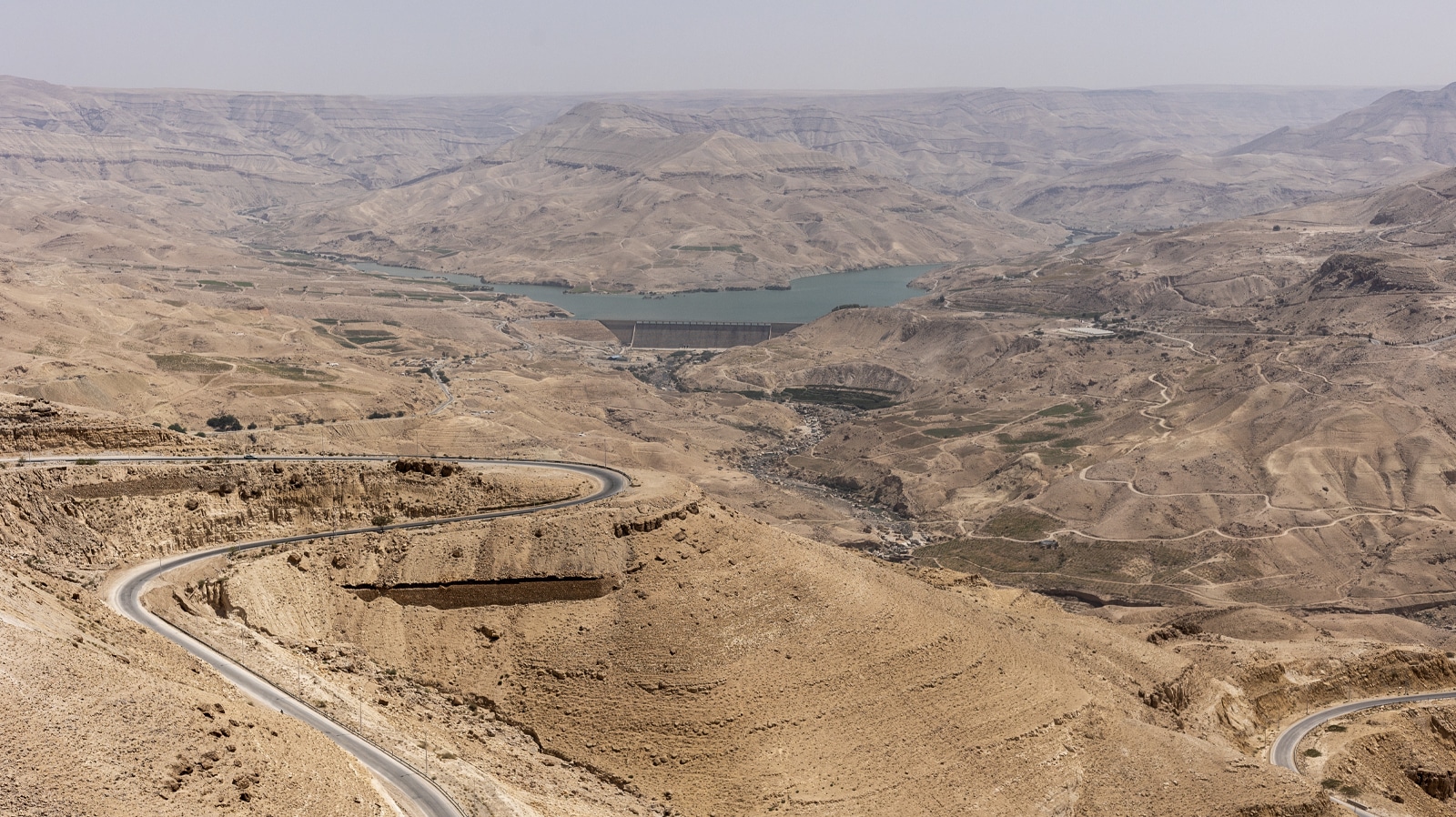A serpentine mountain road descends through arid desert hills toward a turquoise reservoir behind a dam in central Jordan. The rugged canyon landscape stretches under a hazy sky.